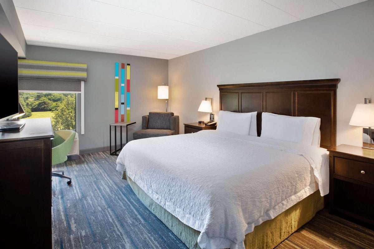 A hotel room with a large bed, a desk with a computer, a chair, a lamp, and a view of greenery outside.