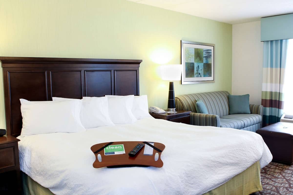 A hotel room with a large bed, a bedside table with a lamp, a sofa, and a small table with a remote control.