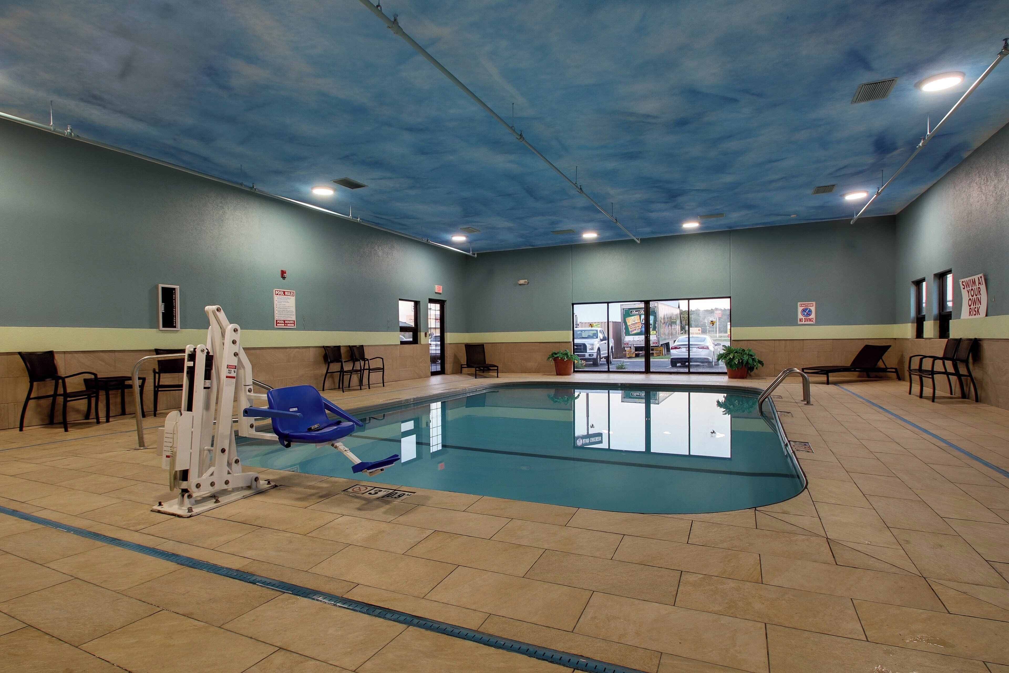 An indoor swimming pool with a lifeguard chair, pool rules sign, and a view of the outside through the glass doors.