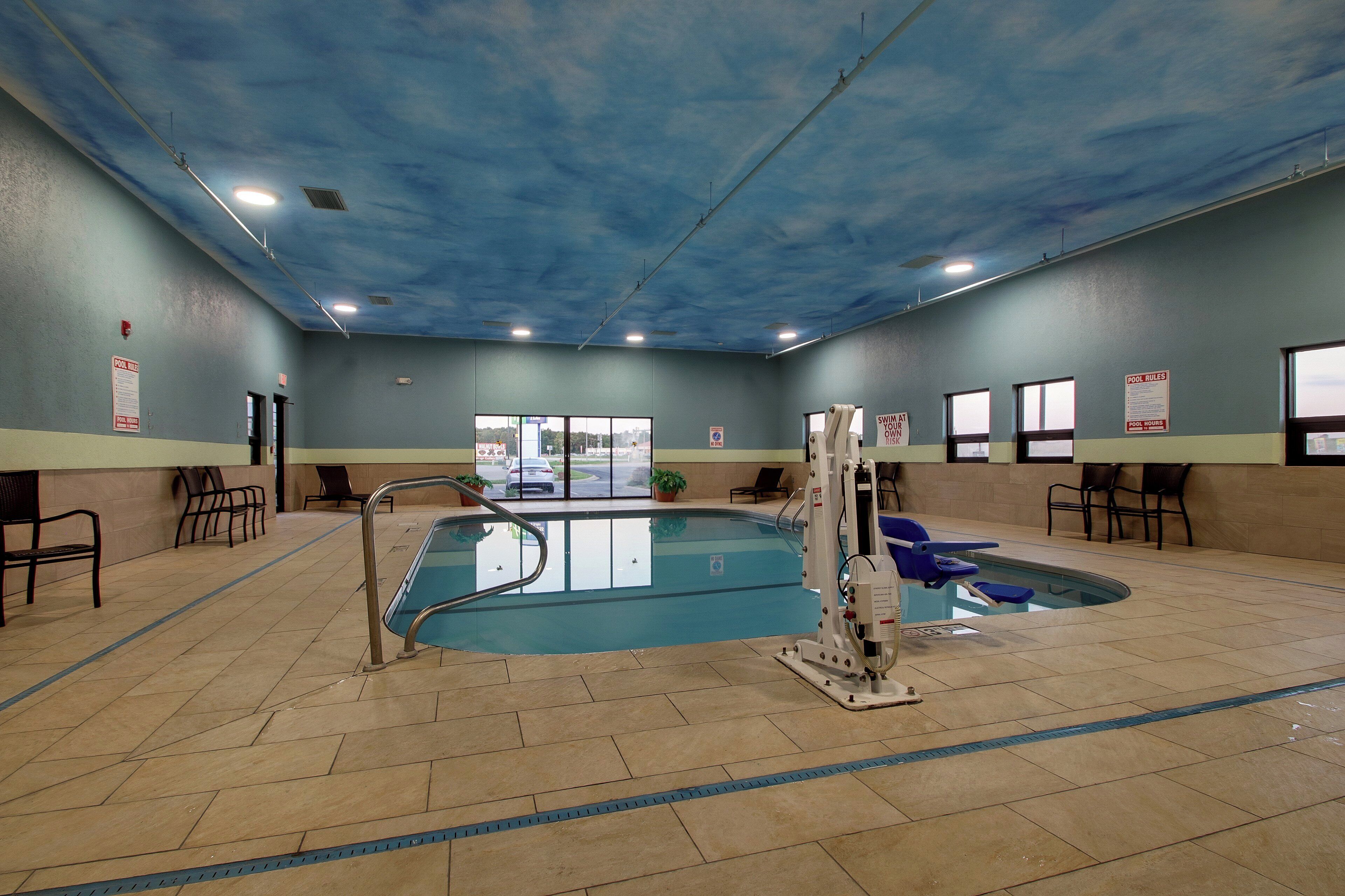 An indoor swimming pool with clear pool rules signs, a lifeguard chair, and a view of the outside through large windows.