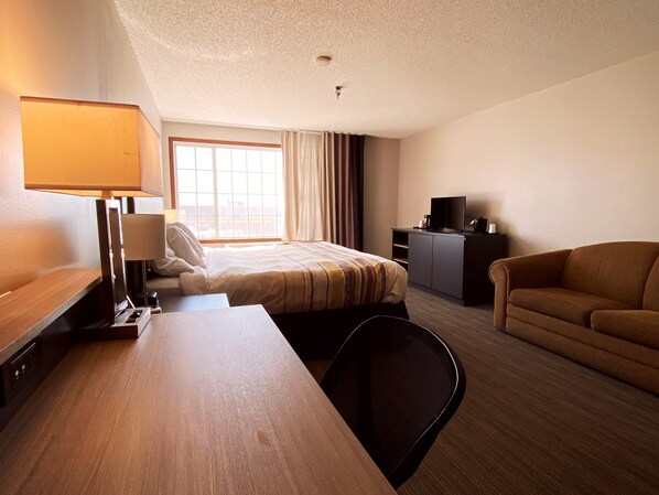 A hotel room with a bed, a desk with a lamp, a TV, a sofa, and a window with curtains.