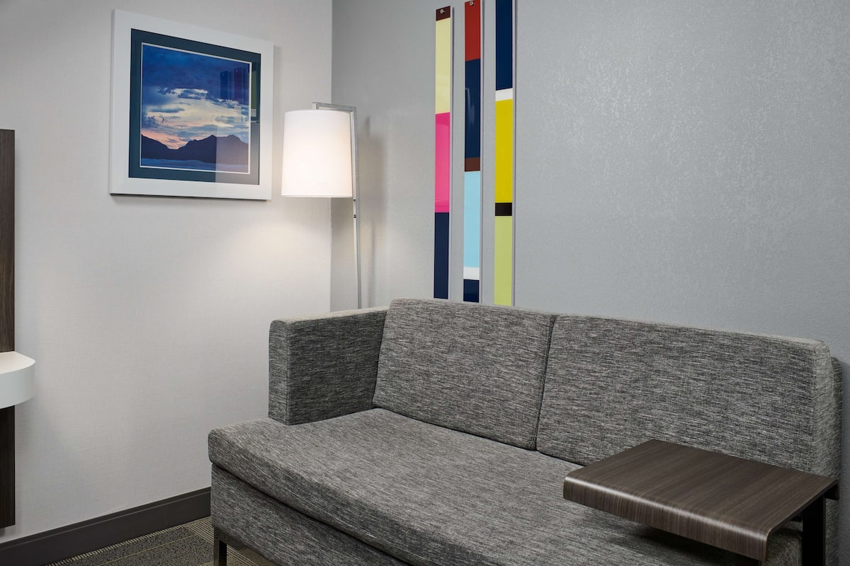 A modern hotel room with a grey sofa, a small wooden side table, a framed picture of a landscape, a floor lamp, and a colorful vertical art piece on the wall.
