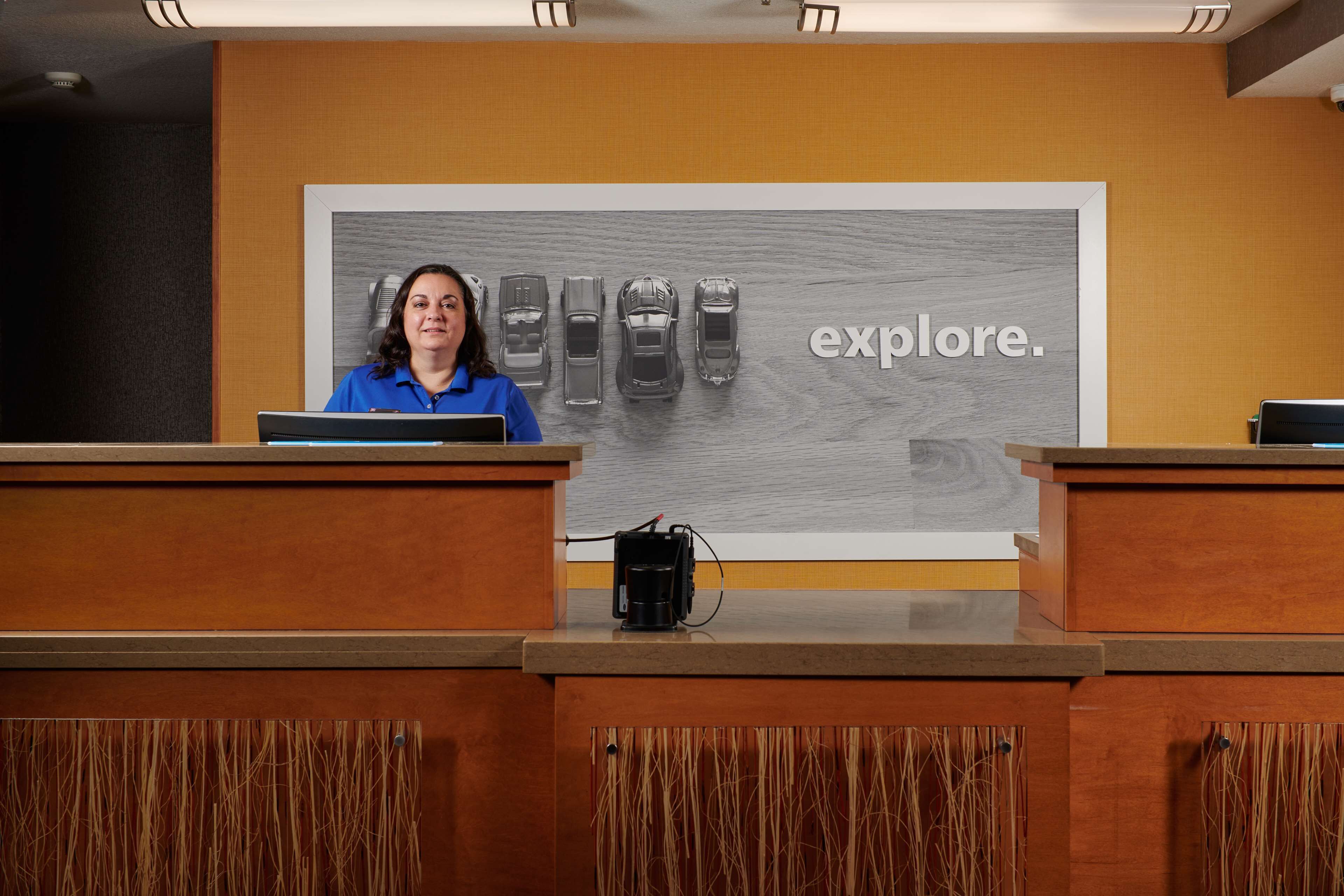 A reception desk with a woman in a blue shirt, a large 'explore.' sign, and a row of cars in the background.