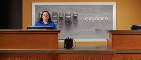 A reception desk with a woman in a blue shirt, a large 'explore.' sign, and a row of cars in the background.