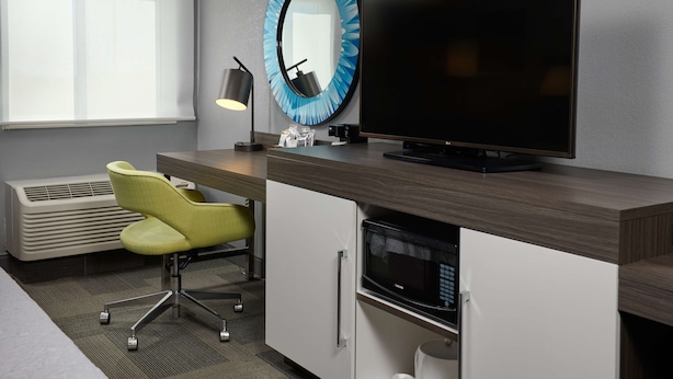 Une chambre d’hôtel moderne équipée d’un téléviseur à écran plat, d’un miroir rond, d’un bureau avec une lampe, d’un four à micro-ondes et d’une chaise.