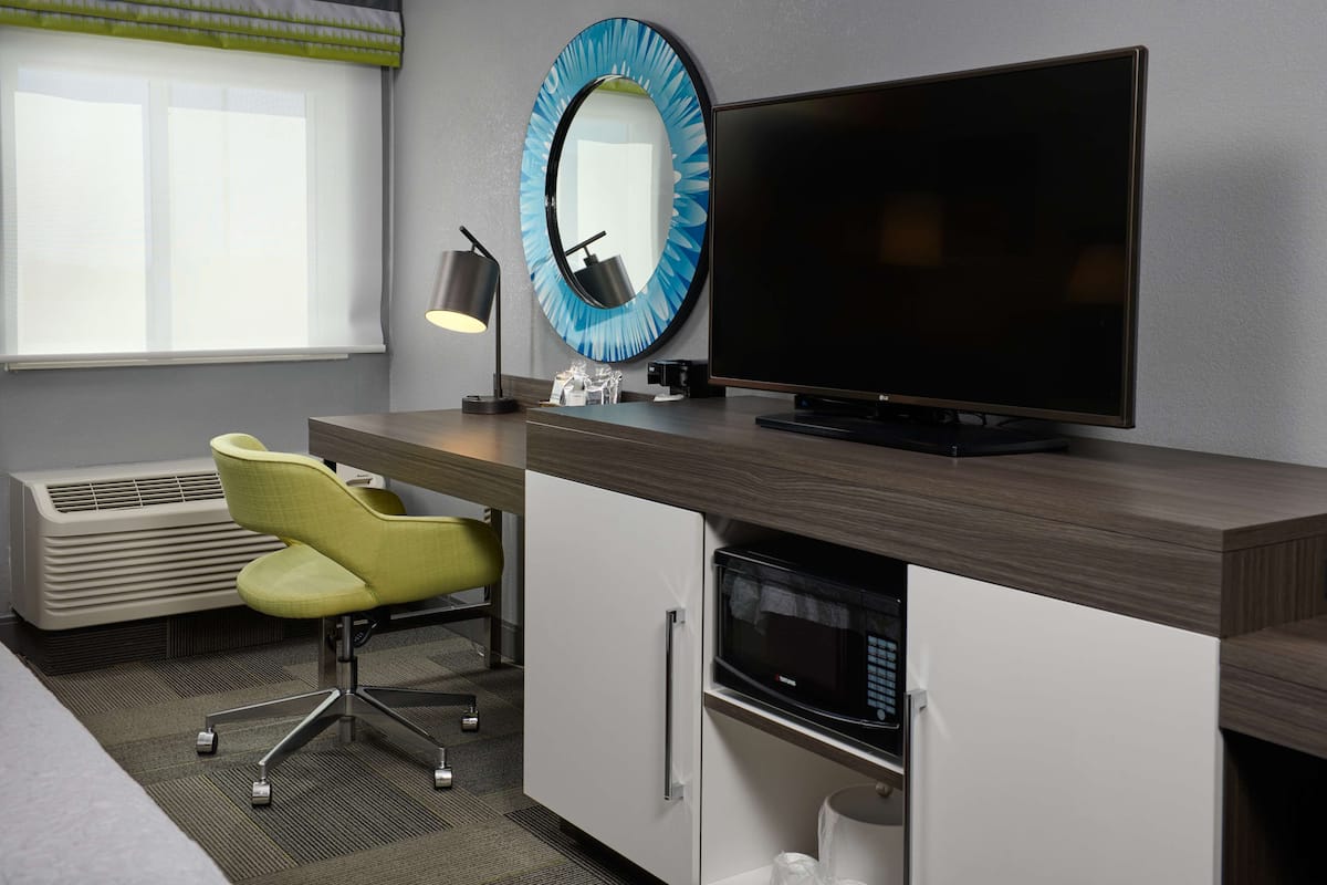 A modern hotel room with a flat-screen TV, a round mirror, a desk with a lamp, a microwave, and a chair.