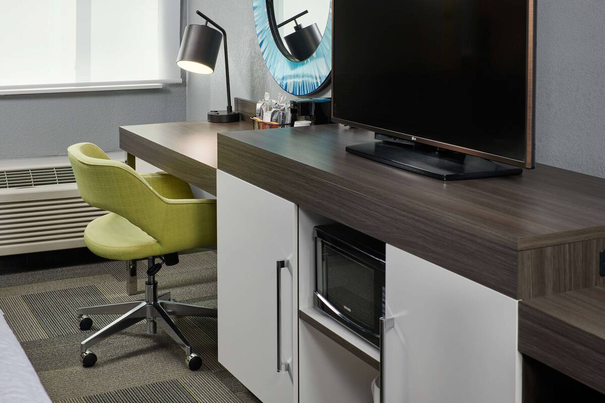 A modern hotel room with a wooden desk, a flat-screen TV, a microwave, and a yellow chair.