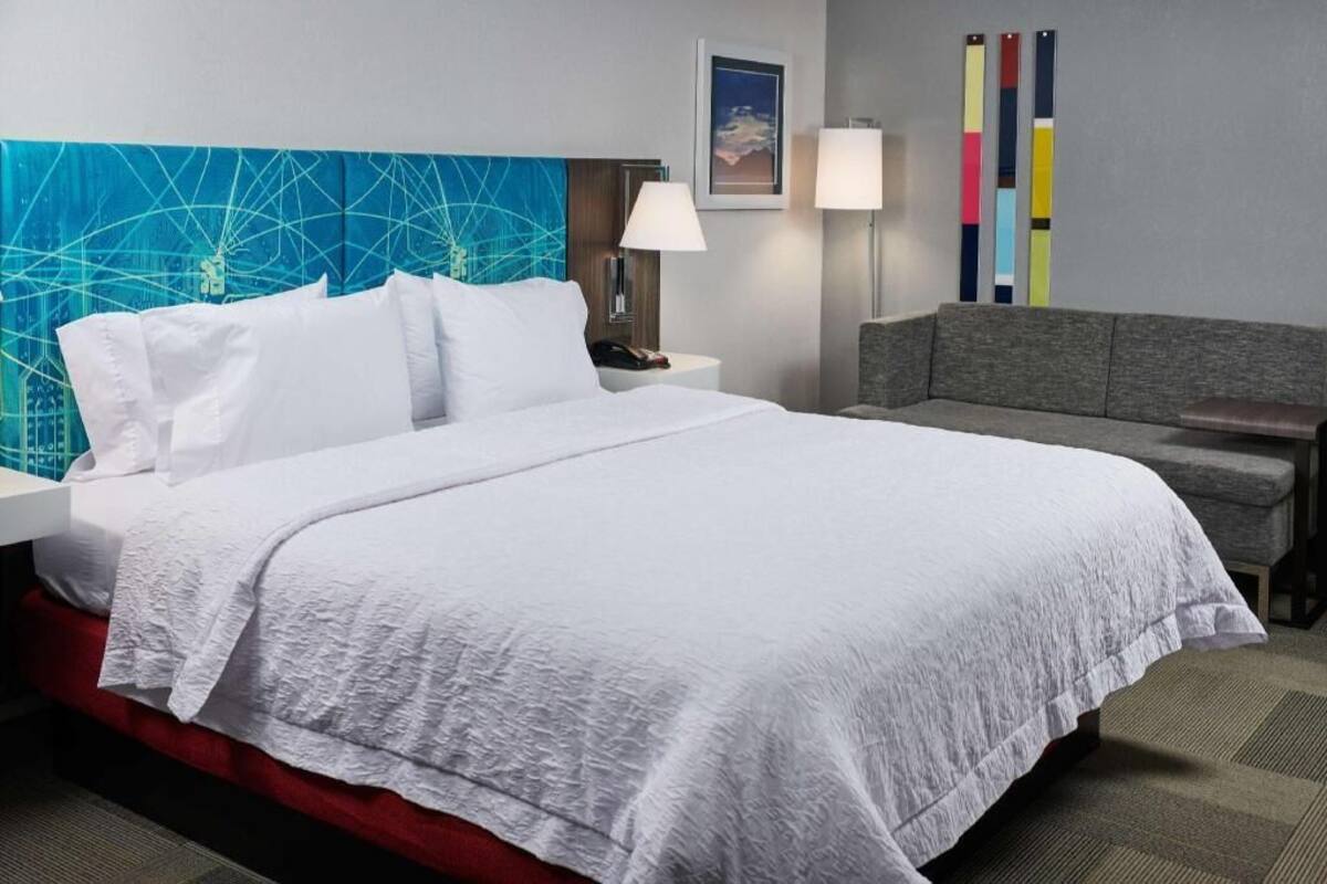 A hotel room with a large bed, a red accent wall, a grey sofa, and a wall with colorful vertical stripes.
