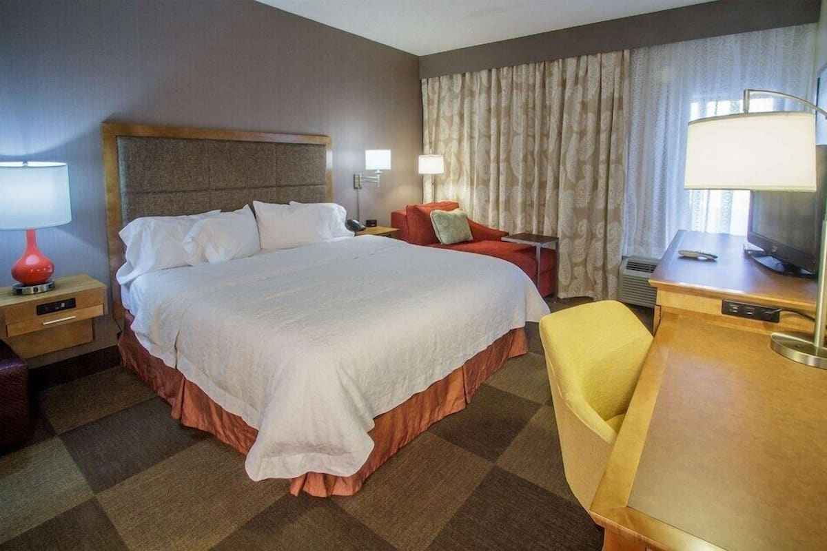 A hotel room with a large bed, bedside tables, a desk with a chair, a television, and a window with curtains.