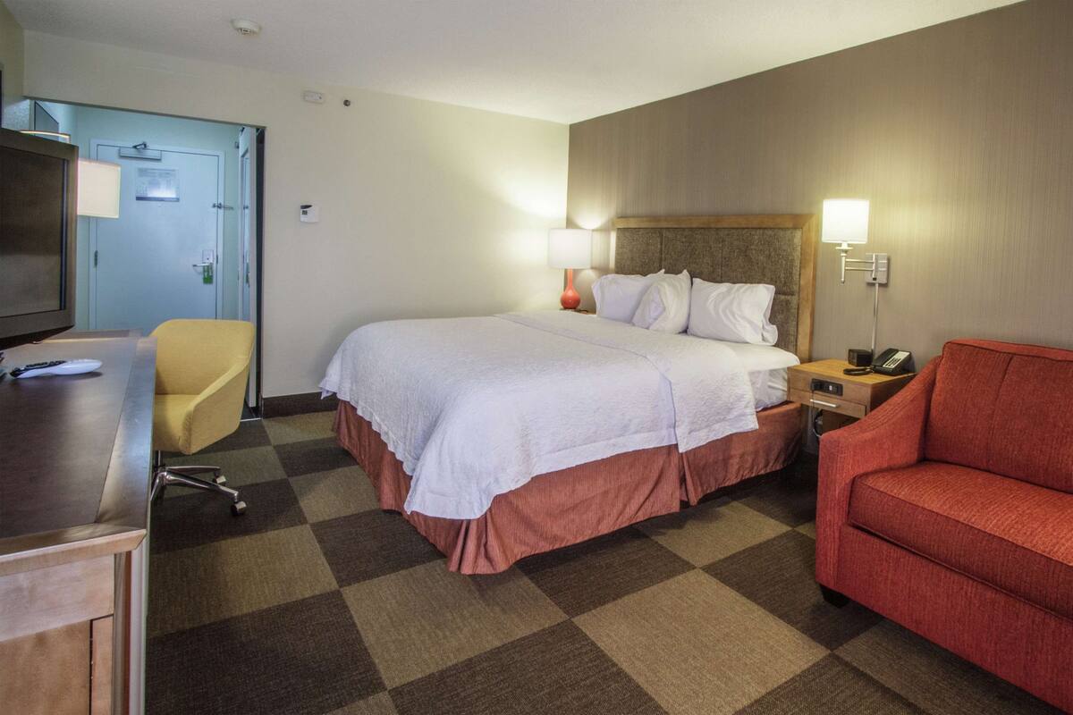 A hotel room with a bed, a red sofa, a desk with a TV and remote, a lamp, and a phone on the nightstand.