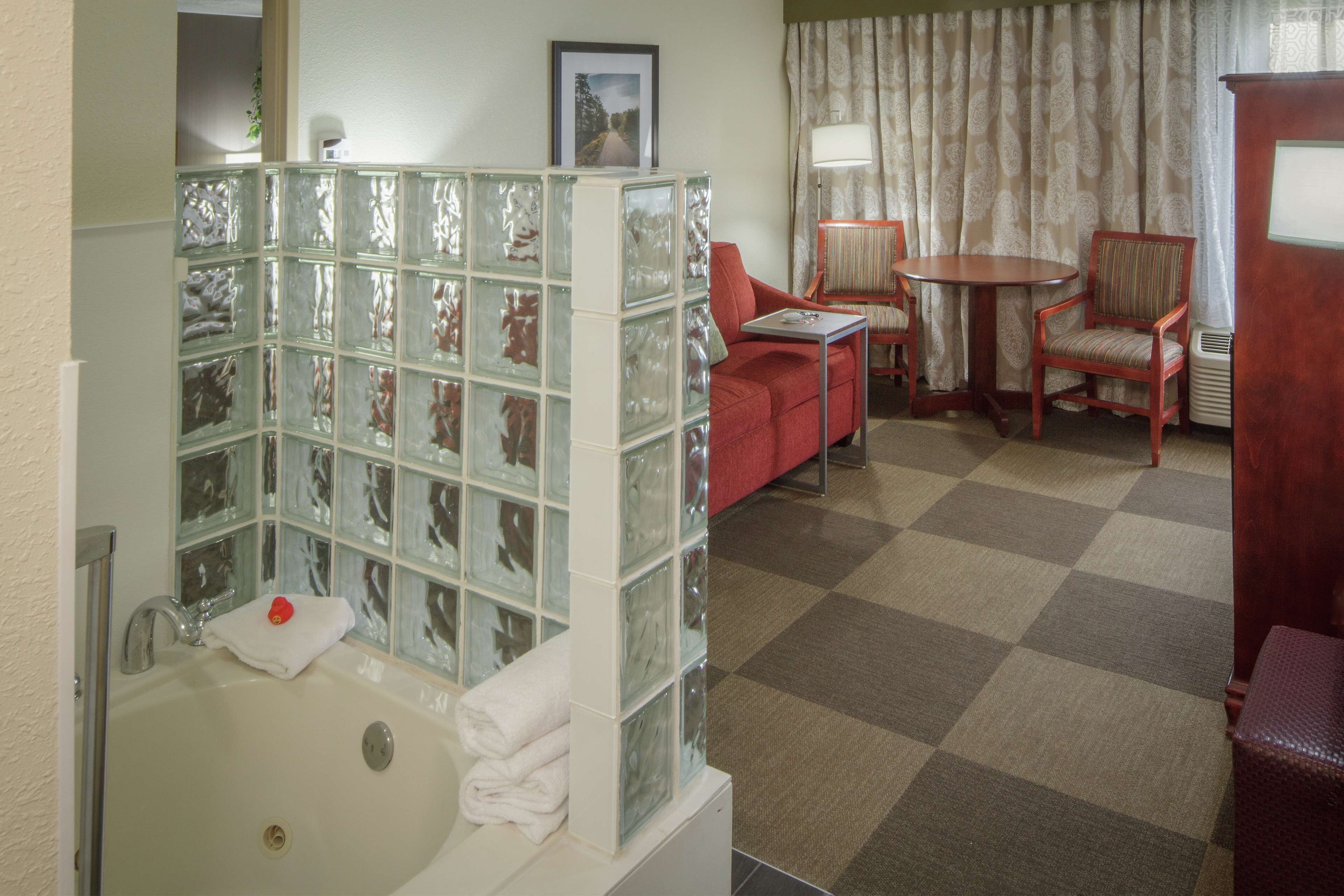 A bathroom with a glass block partition, a bathtub, a red chair, a small table, and a patterned curtain.
