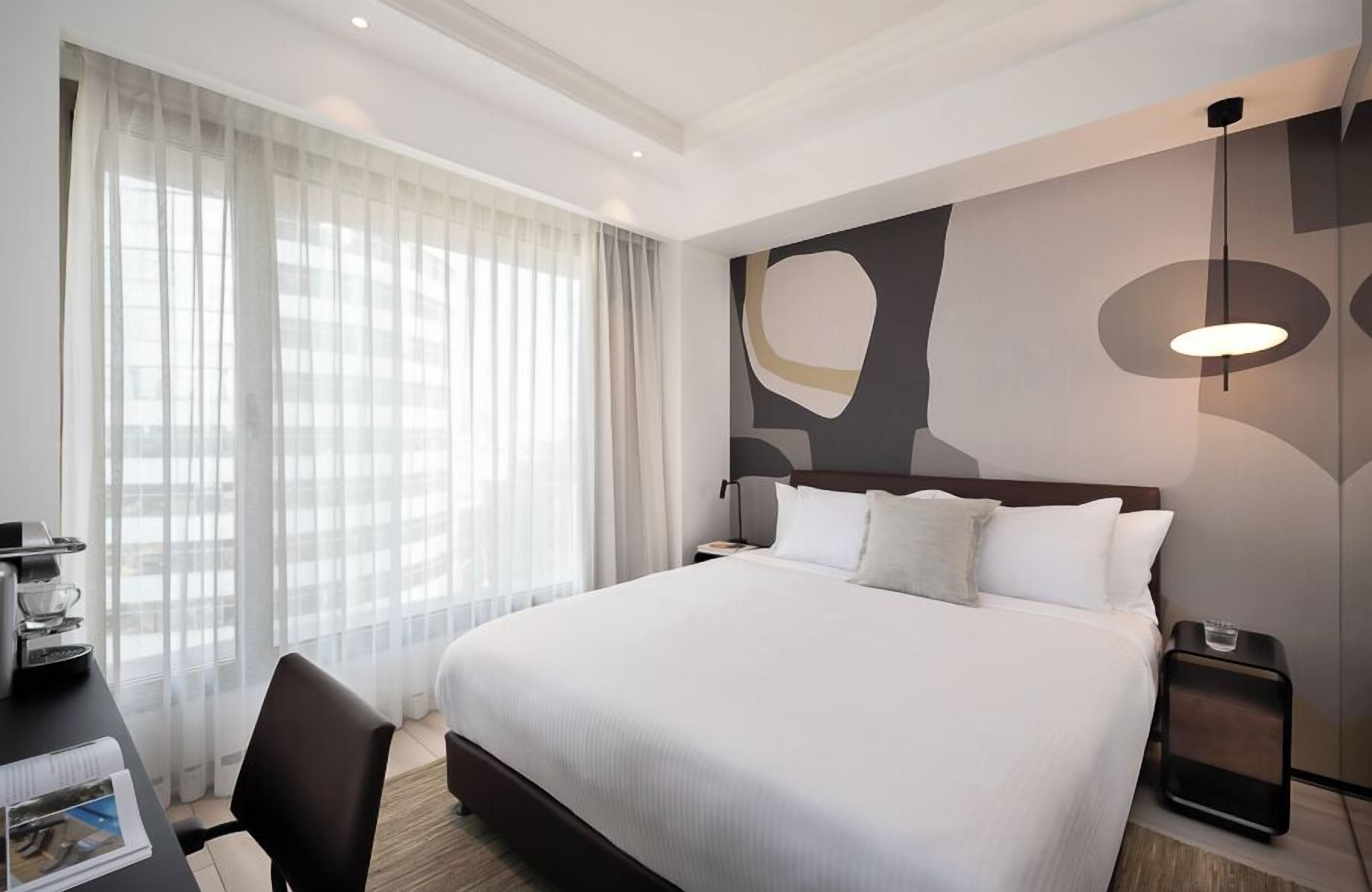 Une chambre d’hôtel avec un grand lit, un bureau équipé d’une machine à café, une chaise et une vue sur la ville à travers de fines rideaux.