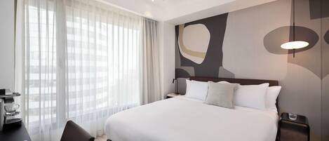 Une chambre d’hôtel avec un grand lit, un bureau équipé d’une machine à café, une chaise et une vue sur la ville à travers de fines rideaux.