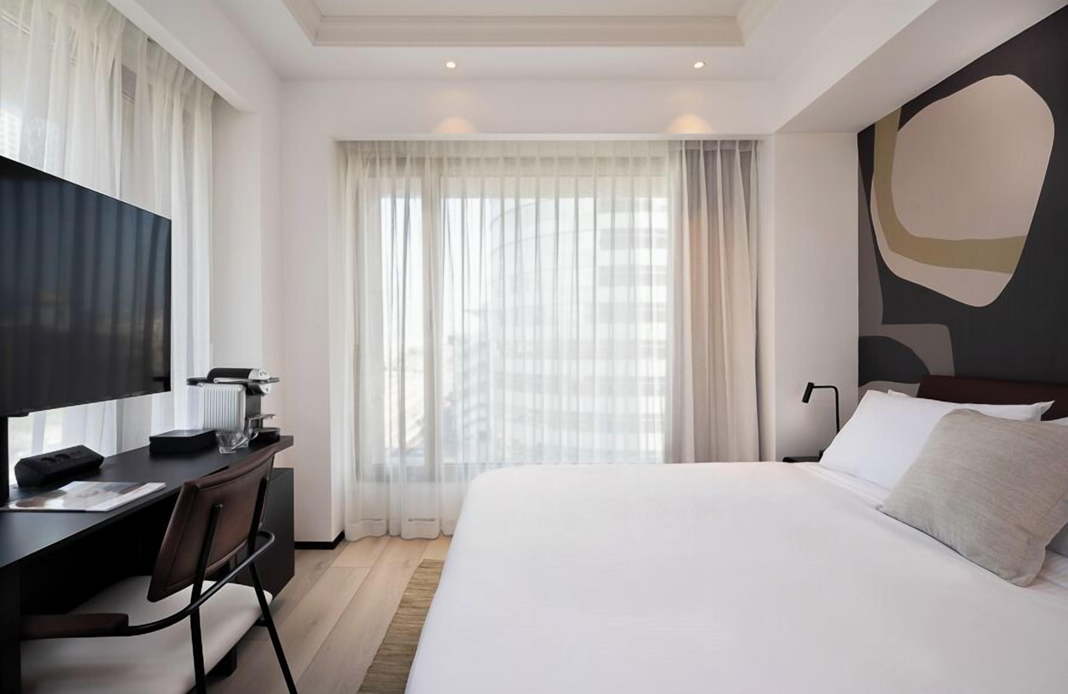 A modern hotel room with a large bed, a desk with a chair, a flat-screen TV, and a large window with sheer curtains.