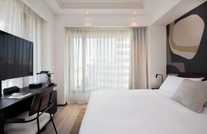Une chambre d’hôtel moderne dotée d’un grand lit, d’un bureau avec une chaise, d’un téléviseur à écran plat et d’une grande fenêtre avec des rideaux légers.
