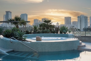 Une piscine sur le toit, avec un jacuzzi, entourée de palmiers et offrant une vue sur les toits de la ville en arrière-plan.