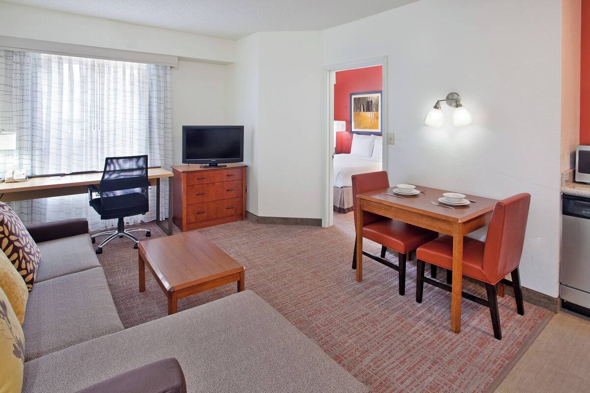 Ein Hotelzimmer mit Sofa, einem kleinen Holztisch, einem Fernseher, einem Schreibtisch und einem Essbereich mit Stühlen.