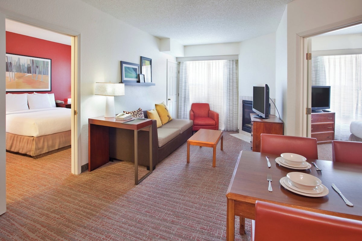 Ein Hotelzimmer mit Bett, Sofa, Esstisch gedeckt mit Tellern und Besteck, Fernseher und einem roten Sessel.