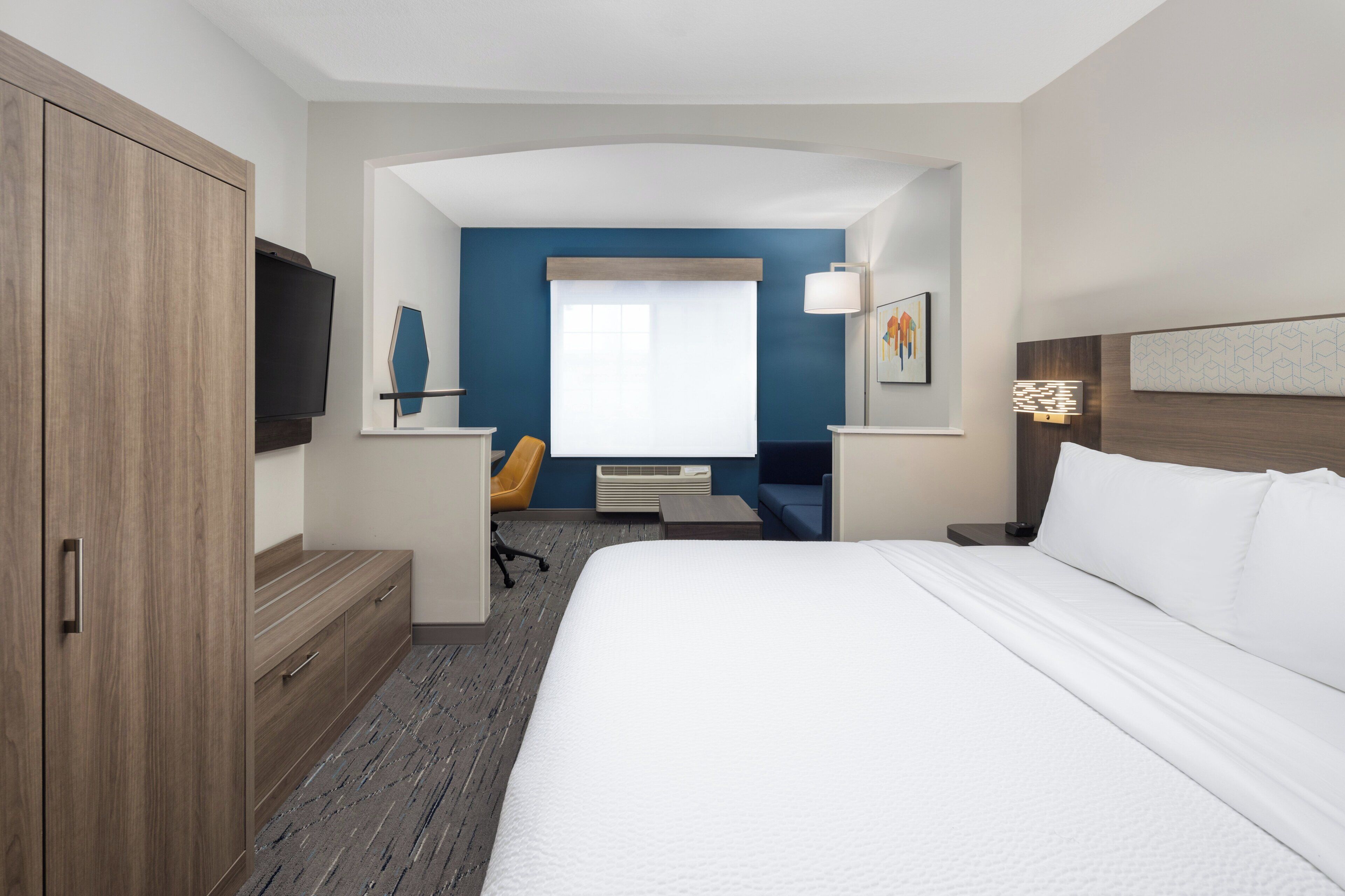 A hotel room with a large bed, a desk with a chair, a flat-screen TV, and a window with curtains.