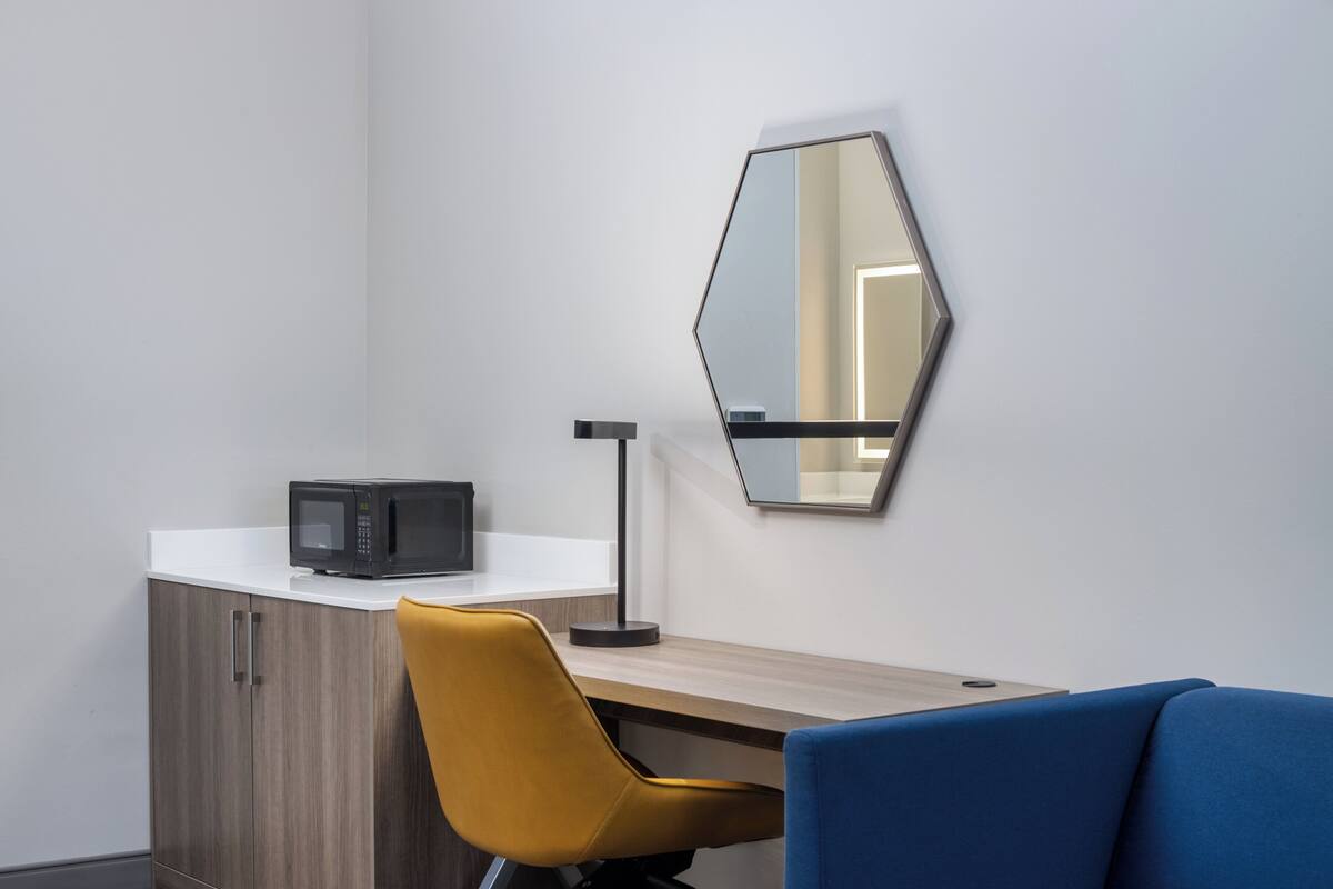 A modern office space with a wooden desk, a yellow chair, a blue chair, a black microwave, and a large mirror on the wall.