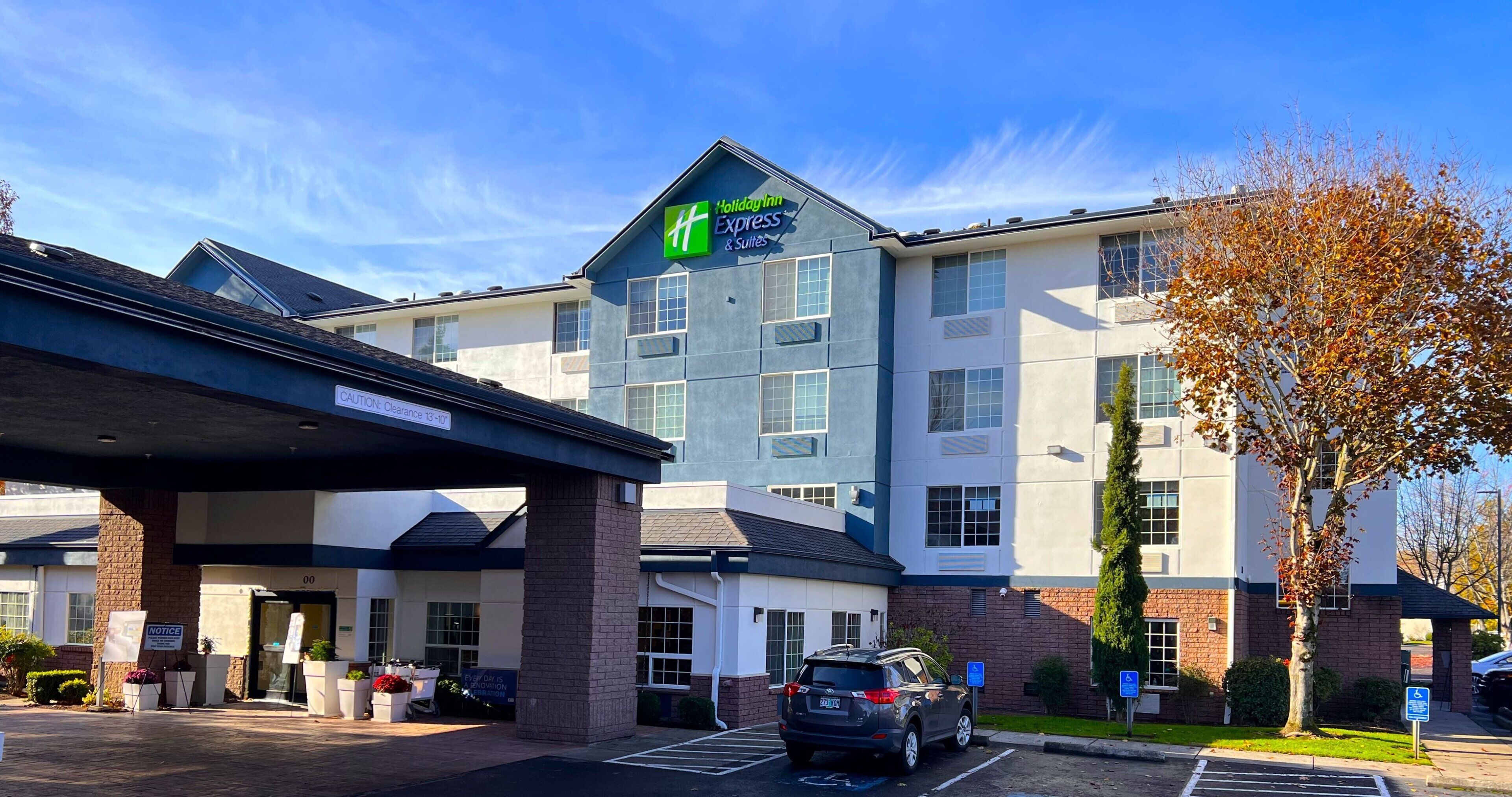 A Holiday Inn Express & Suites building with a clear sky, parking area, and a tree with autumn leaves.