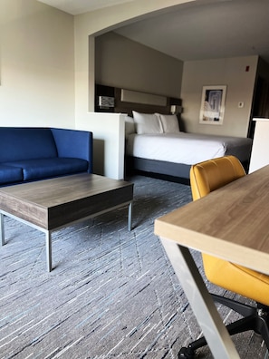 A hotel room with a blue sofa, a wooden coffee table, a yellow chair, a bed with white bedding, and a framed picture on the wall. - Holiday Inn Express Hotel & Suites Portland-Jantzen Beach by IHG (Portland)
