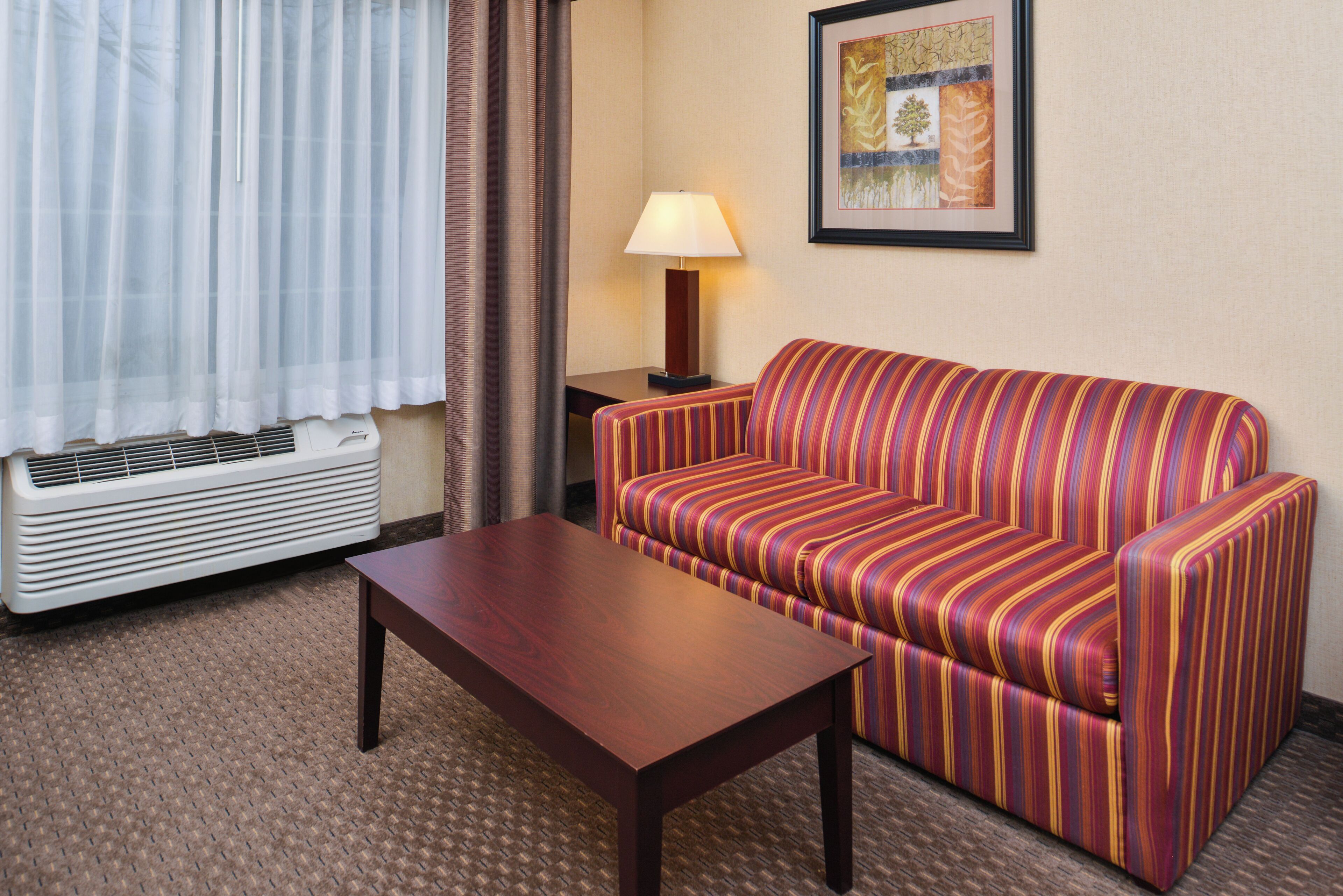 Een hotelkamer met een gestreepte bank, een houten tafel, een lamp en een kunstwerk aan de muur.