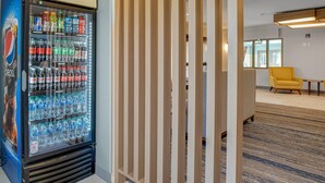 A vending machine stocked with various beverages, a wooden partition, and a yellow armchair in a modern interior. - Holiday Inn Express Hotel & Suites Portland-Jantzen Beach by IHG (Portland)