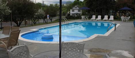 Piscina con sillones de descanso y una mesa, rodeada por una valla y árboles.