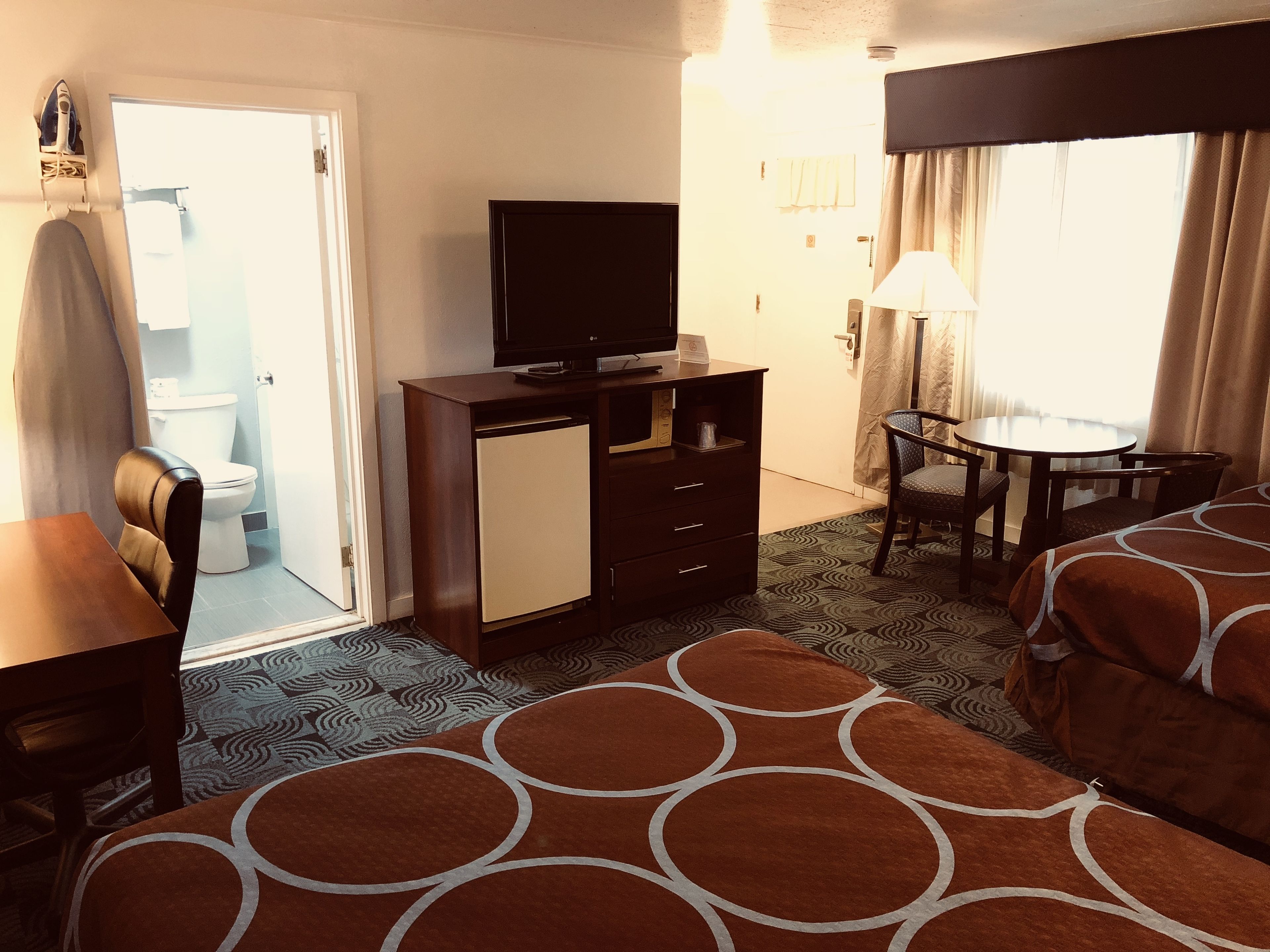 A hotel room with a bed, a desk, a chair, a television, a small cabinet, a bathroom visible through an open door, and a patterned rug.