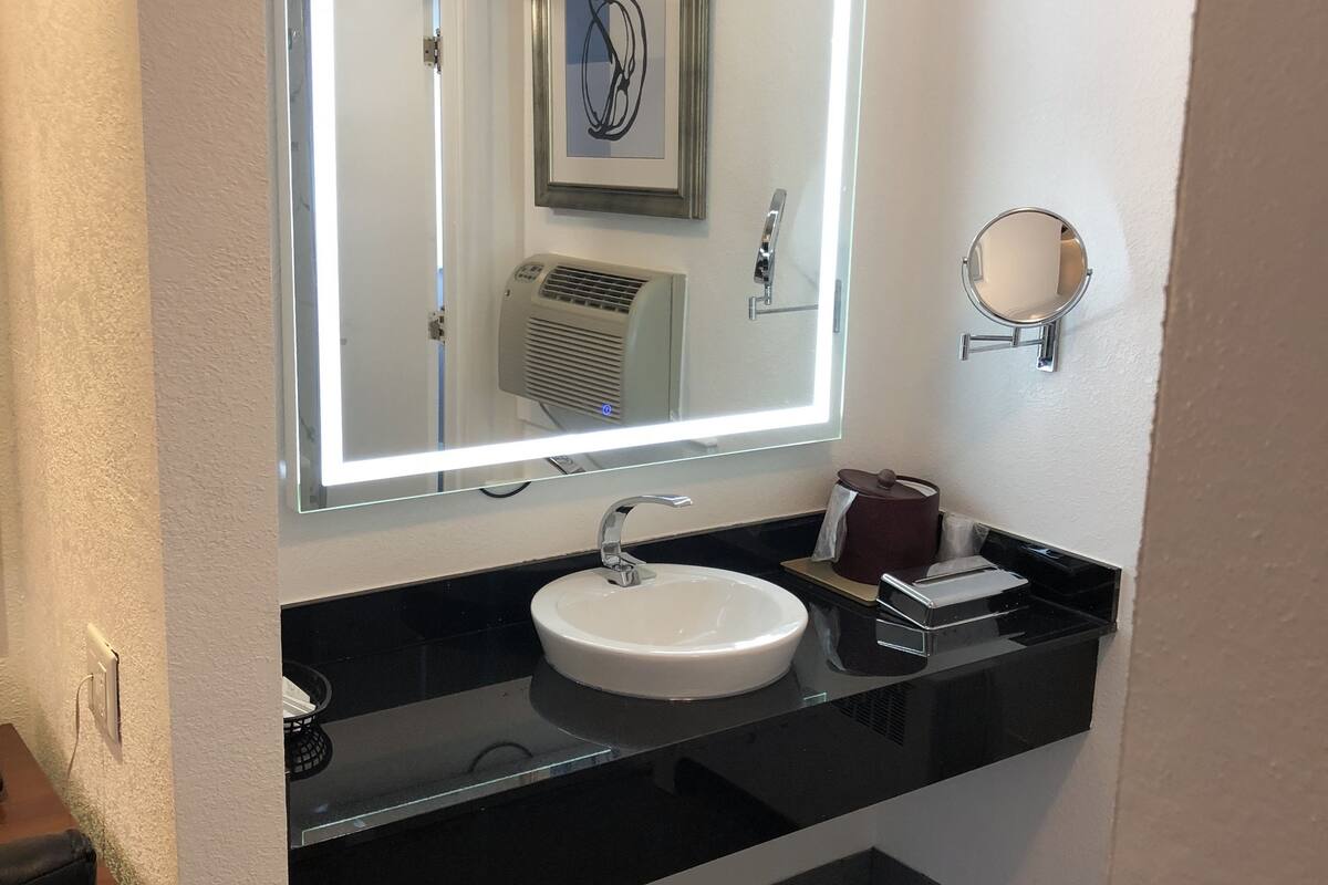 A bathroom with a white sink, a mirror with a light, a heater, and a trash can.