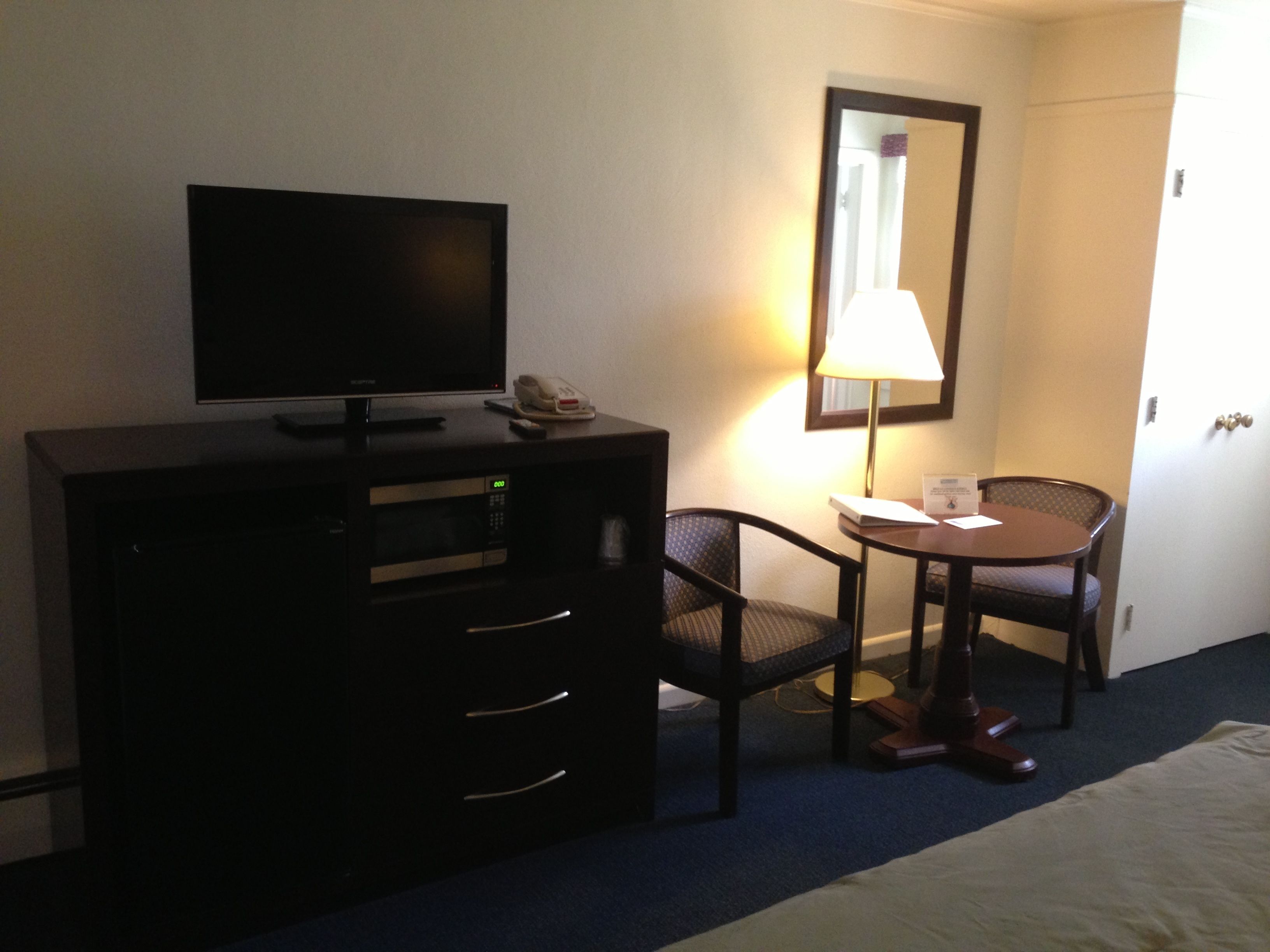 A hotel room with a TV, a desk with a lamp, a chair, and a bed.