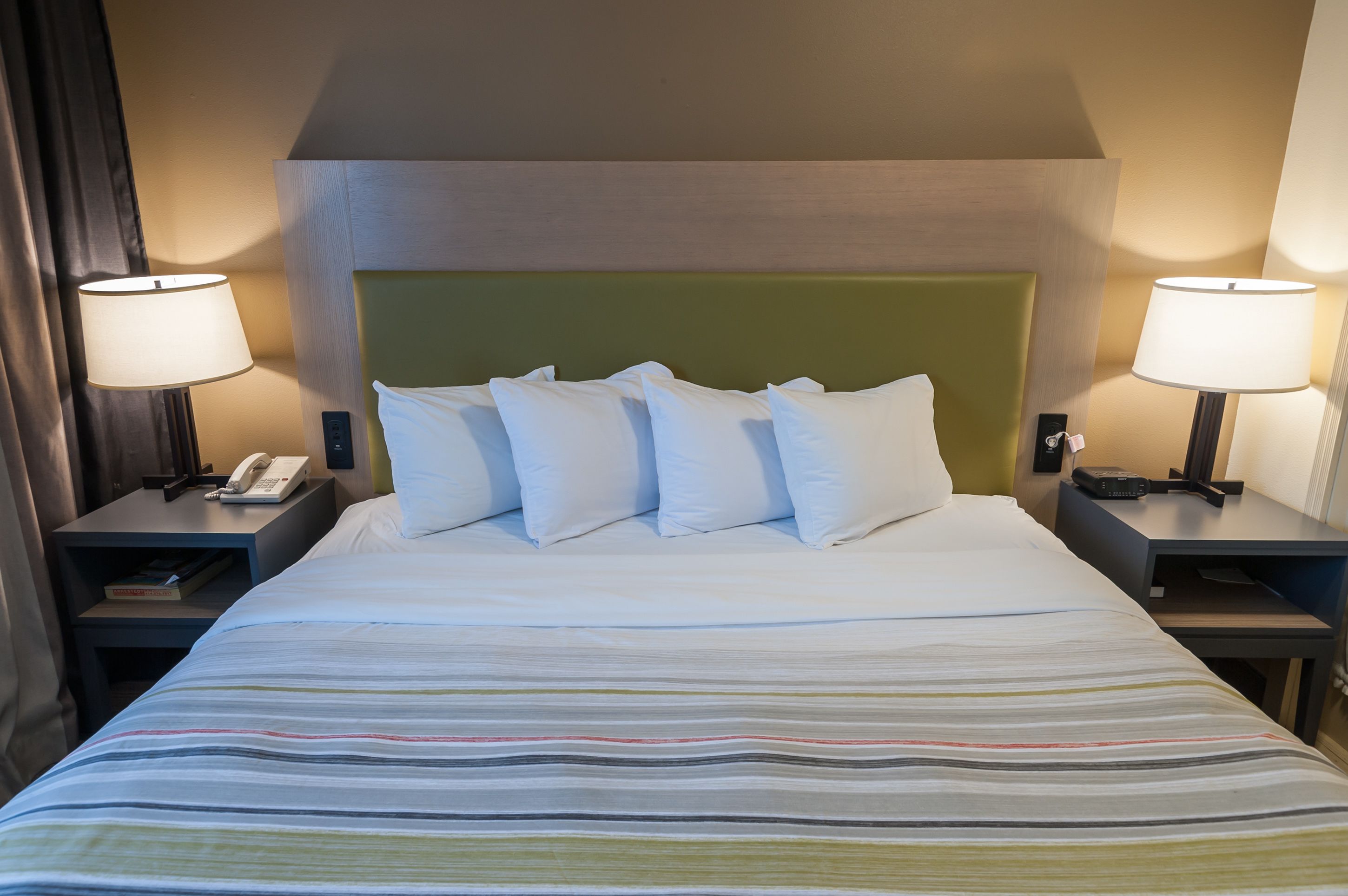 Quarto de hotel com uma cama grande, duas mesas de cabeceira, um painel de cabeceira verde e duas luminárias.