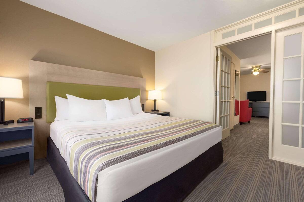 A hotel room with a large bed, two bedside tables with lamps, a headboard, and a door leading to a hallway with a TV and a red chair.