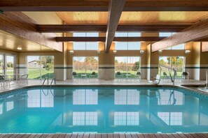 A large indoor swimming pool with clear blue water, wooden beams on the ceiling, and multiple windows offering views of the surrounding area.