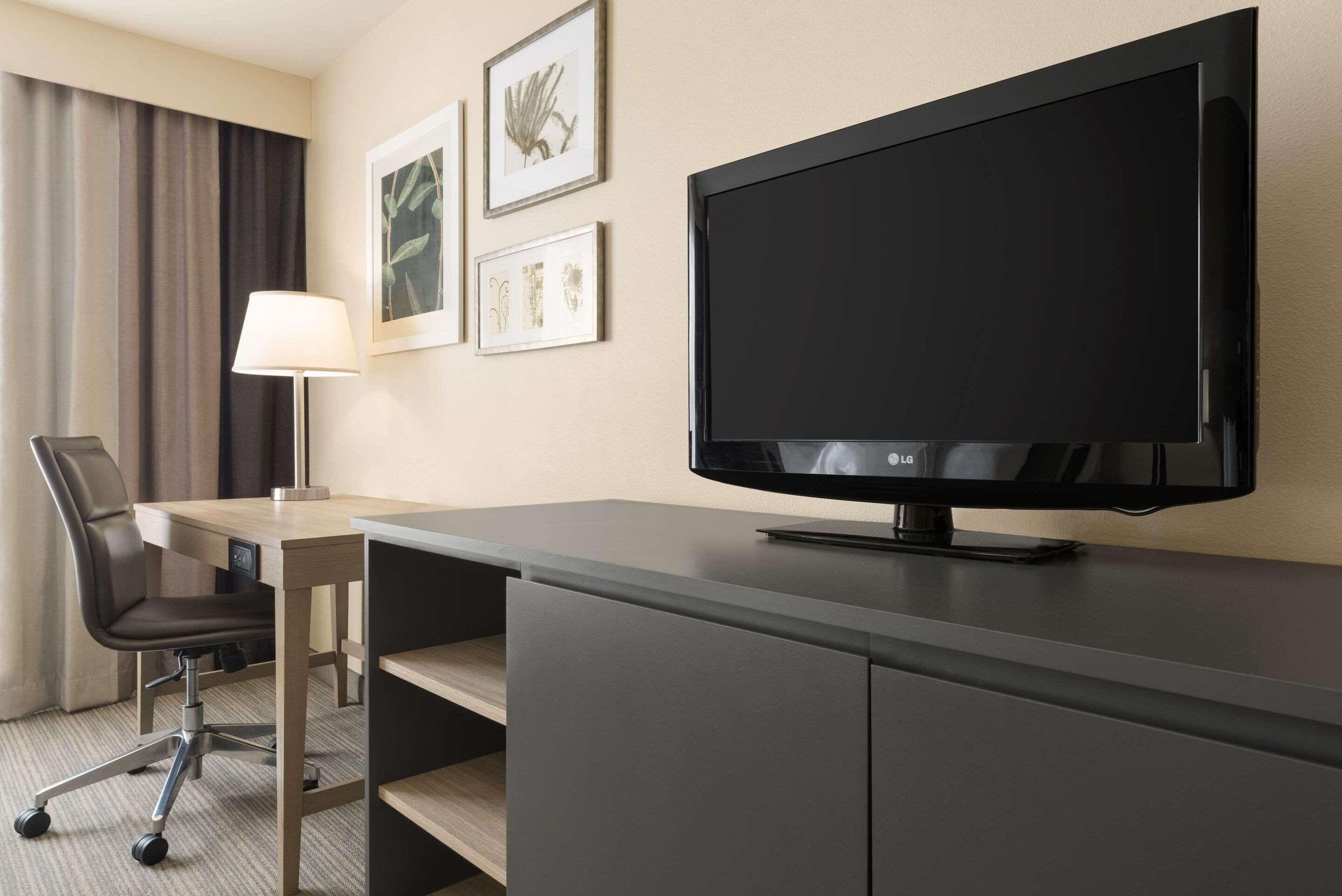 Habitación de hotel con escritorio, silla, televisor, lámpara y una obra de arte enmarcada en la pared.