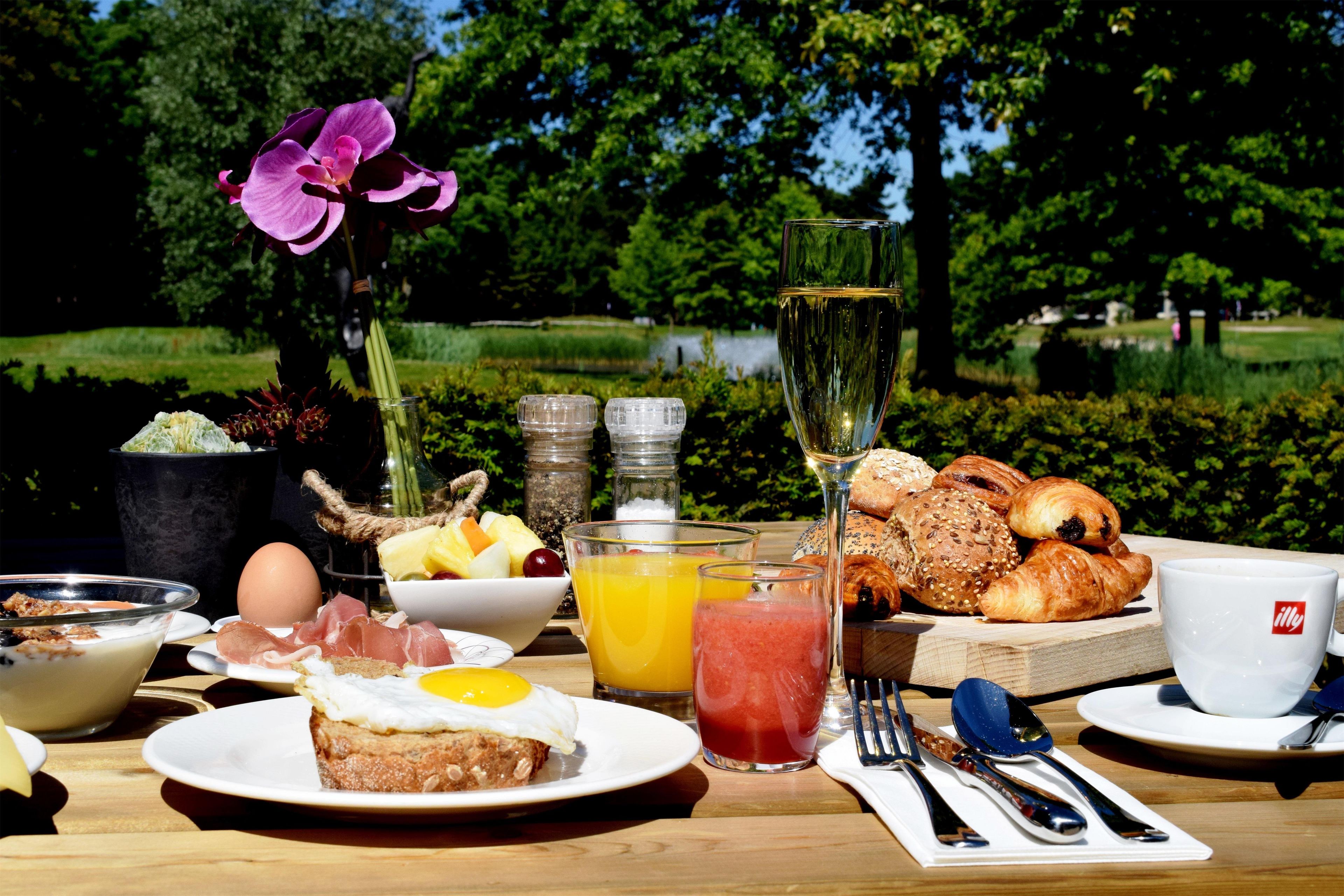 A breakfast table with food and drinks, a flower, and a view of a park.