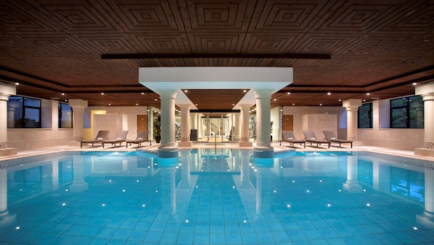 A large indoor swimming pool with clear blue water, surrounded by lounge chairs and columns, and a wooden ceiling with geometric patterns. - DoubleTree by Hilton Royal Parc Soestduinen (Soest)