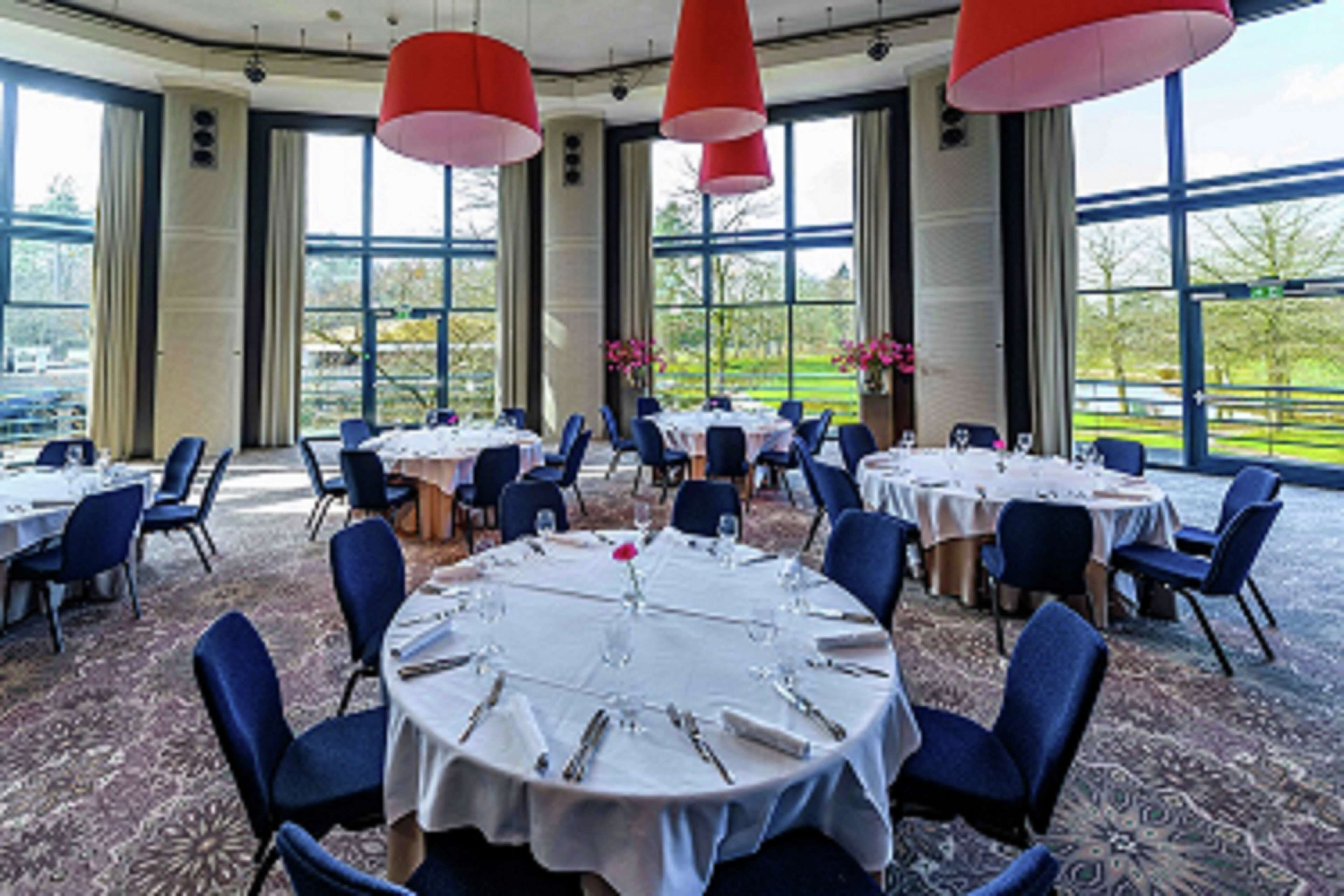 Un salón de banquetes con mesas redondas dispuestas para cenar, ventanas amplias y lámparas colgantes rojas.