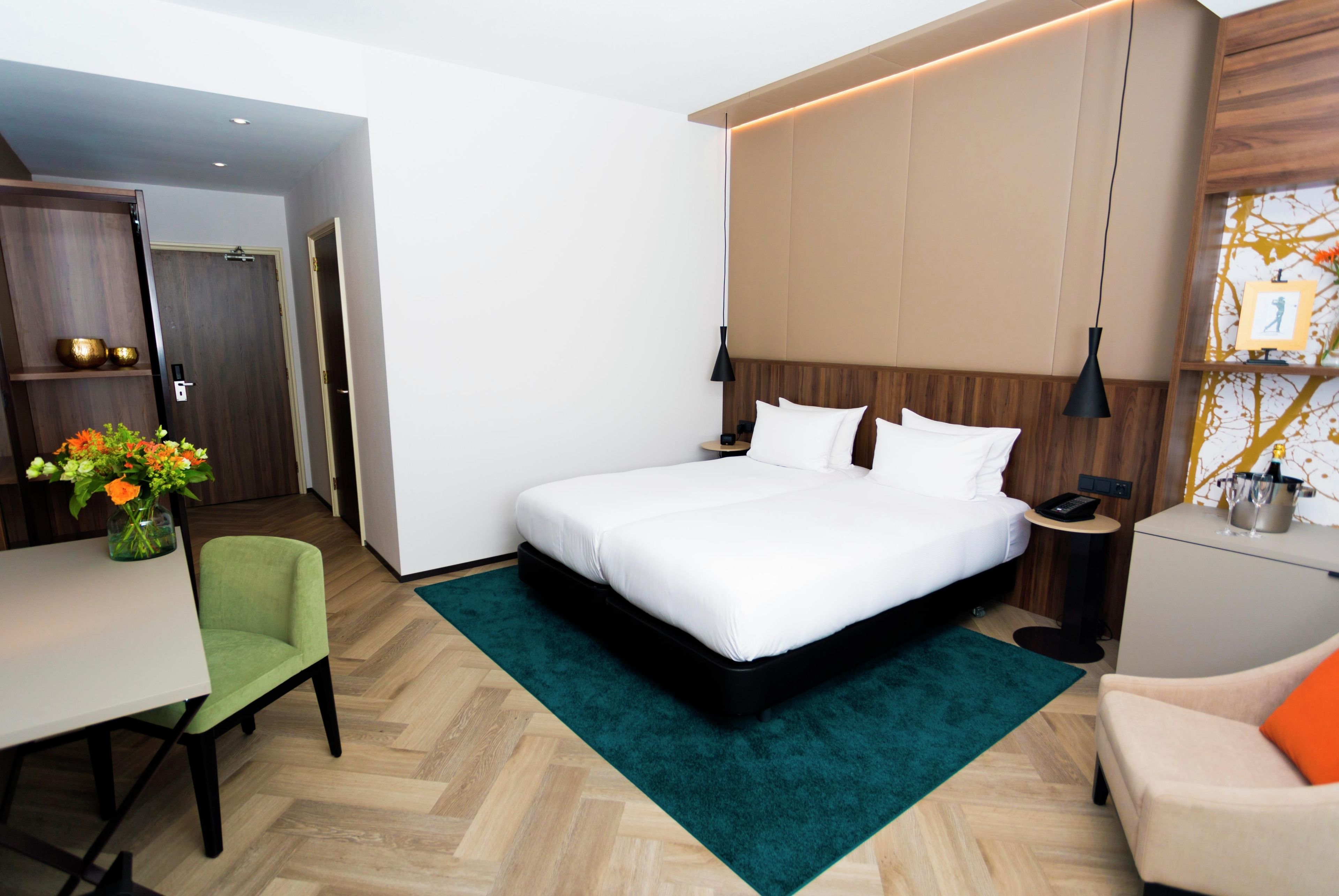 Een hotelkamer met een bed, nachtkastje, lamp, stoel, bureau en een vaas met bloemen.