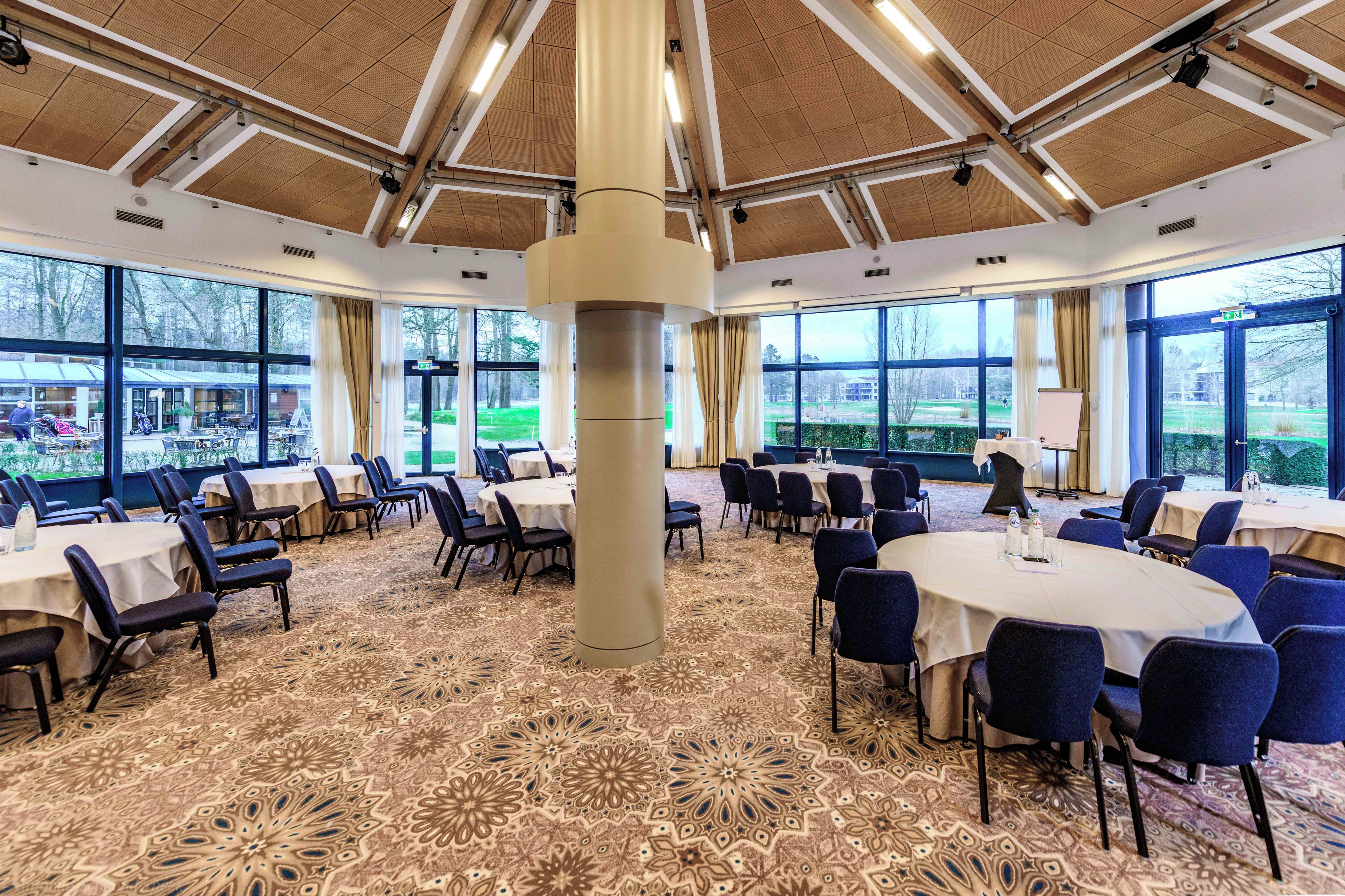 Une salle de conférence dotée de tables et de chaises rondes, de grandes fenêtres et d’un tapis à motifs.