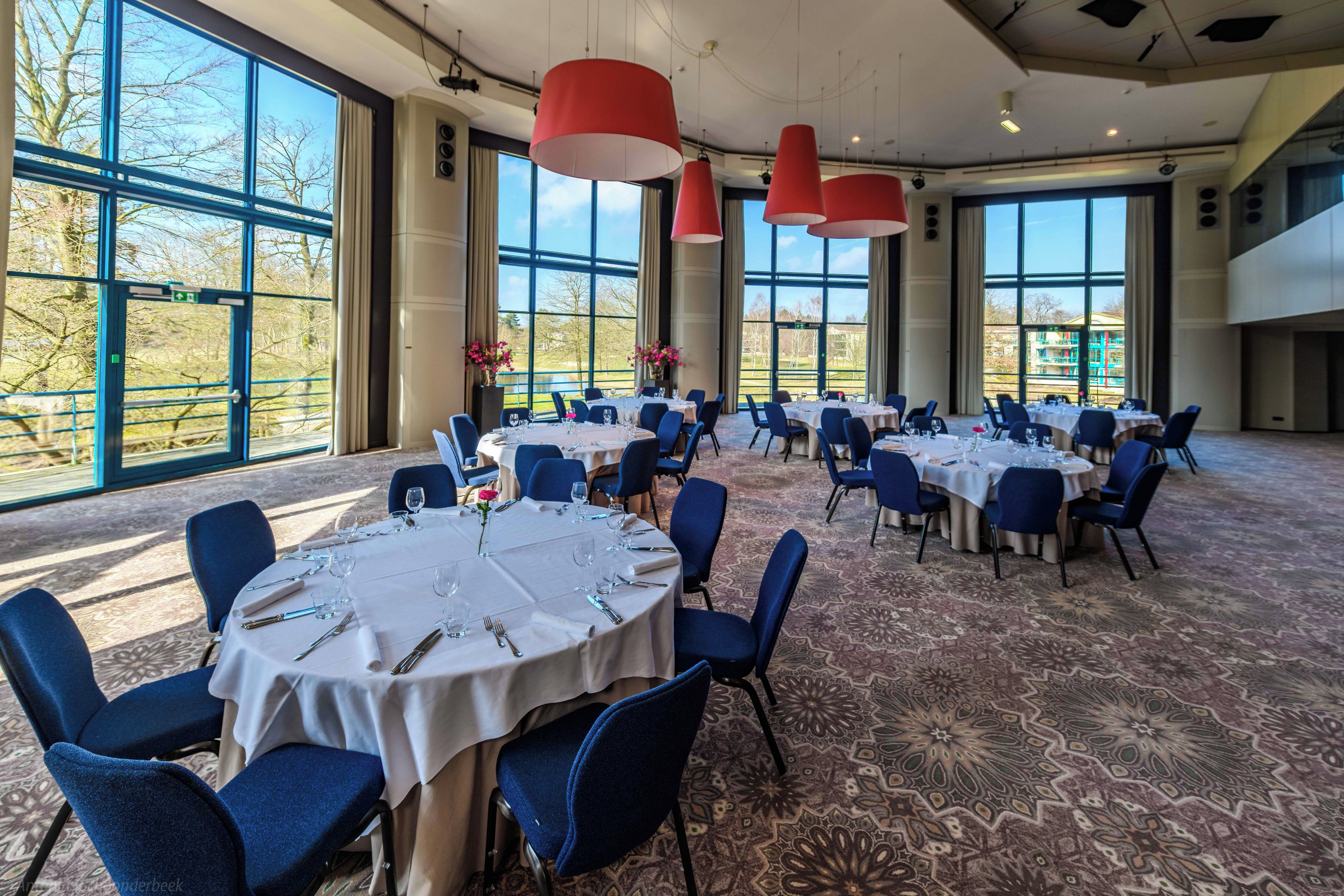 A conference room with round tables, blue chairs, and large windows offering a view of the outdoors.