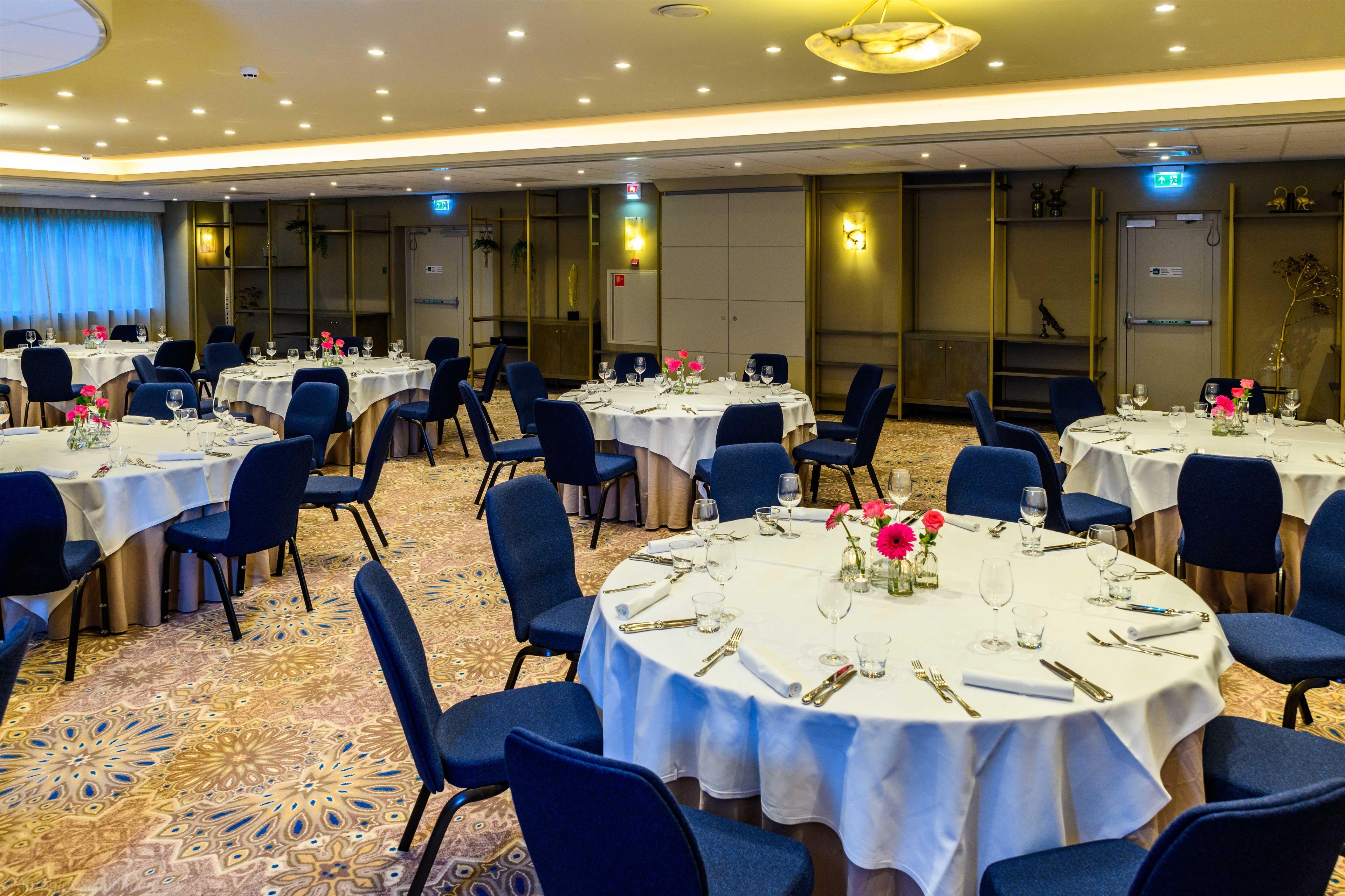Un salón de banquetes con mesas redondas dispuestas para comer, sillas azules y una alfombra estampada.