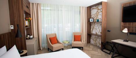 A hotel room with a large bed, two armchairs, a desk, a small table, and a TV.