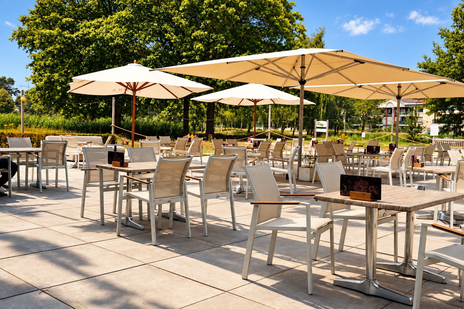 An outdoor café with multiple tables and chairs, shaded by large umbrellas, set against a backdrop of trees and a clear sky.