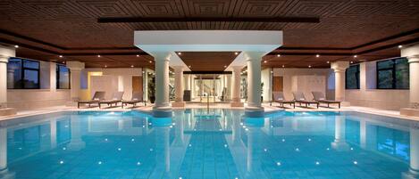A large indoor swimming pool with clear blue water, surrounded by lounge chairs and columns, with a patterned ceiling and large windows.