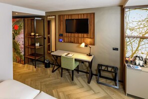 A hotel room with a wooden desk, a television mounted on the wall, a small table with a lamp, and a view of trees through the window.