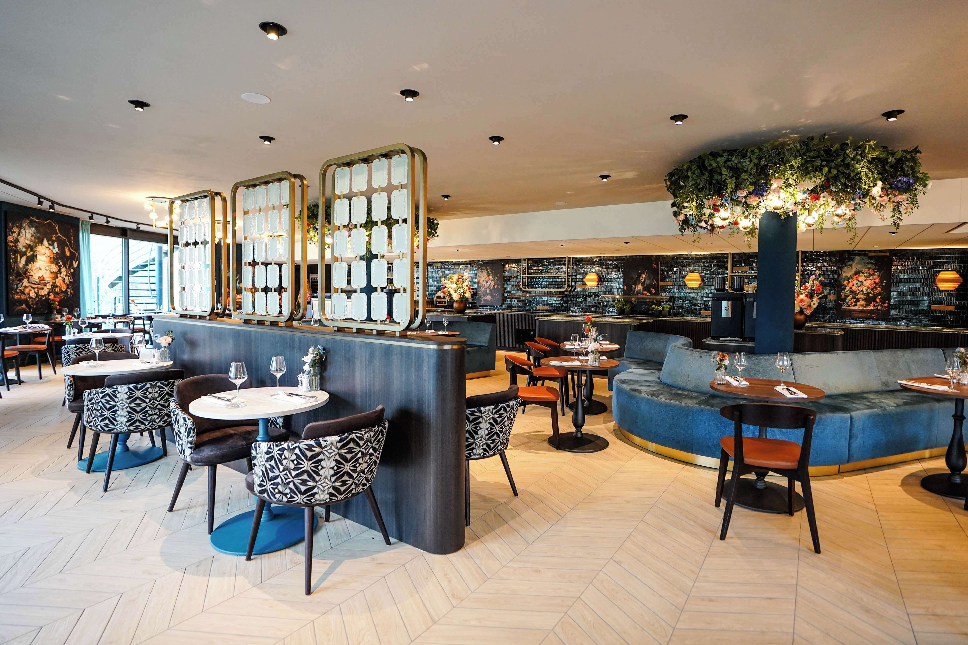 Un restaurante moderno con mesas redondas, sillas estampadas, una barra central y una decoración floral colgante.