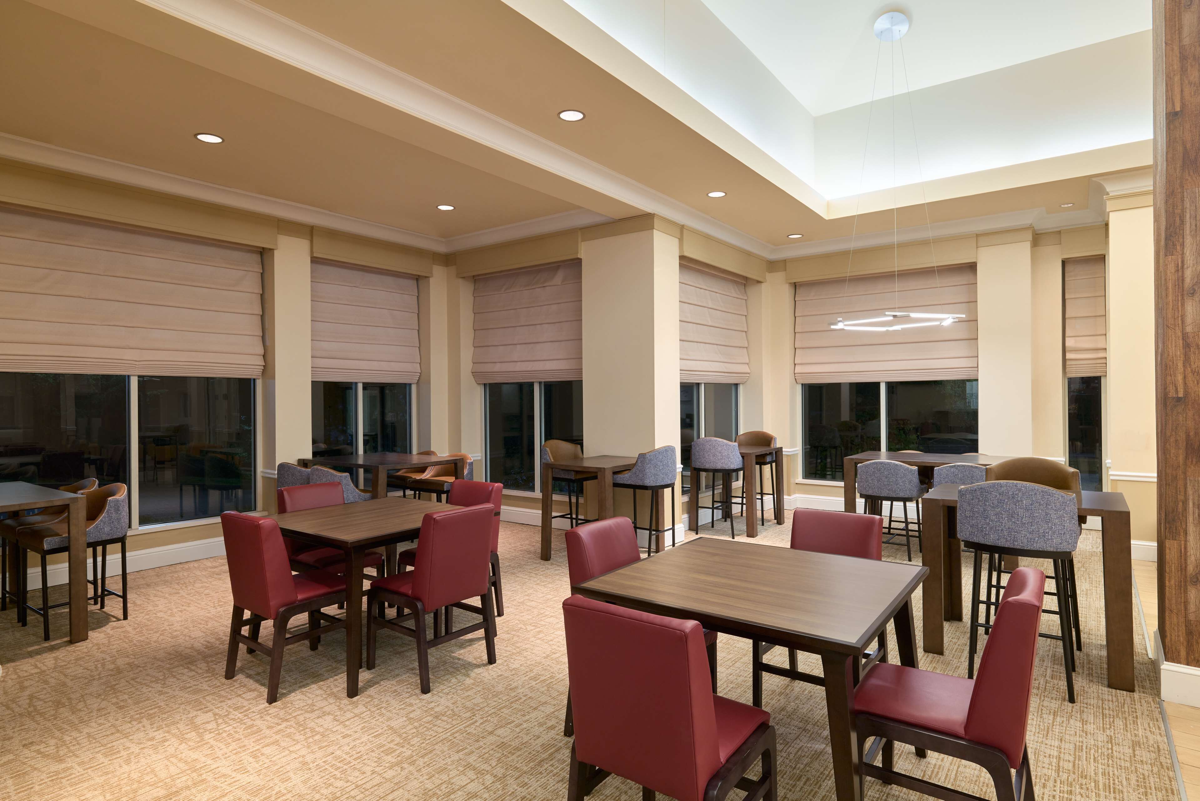 A spacious indoor dining area with multiple tables and chairs, large windows with blinds, and a high ceiling with recessed lighting.
