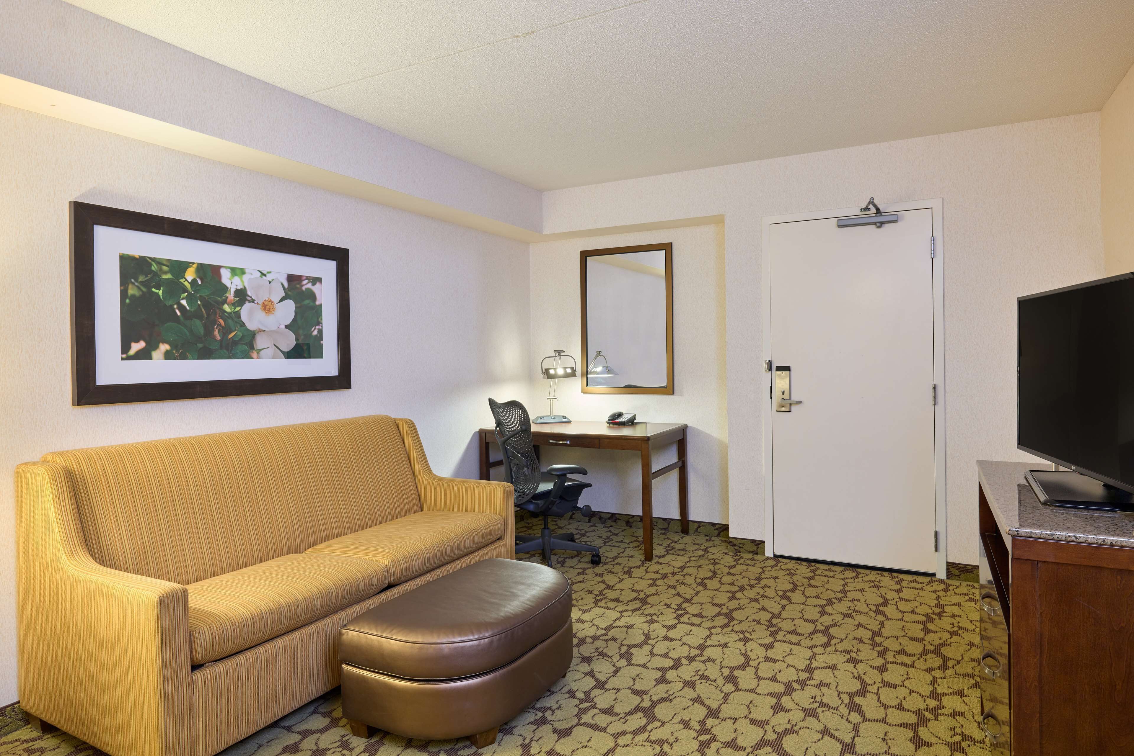 Habitación de hotel con sofá, reposapiés, escritorio con silla, televisión, espejo y un cuadro en la pared.